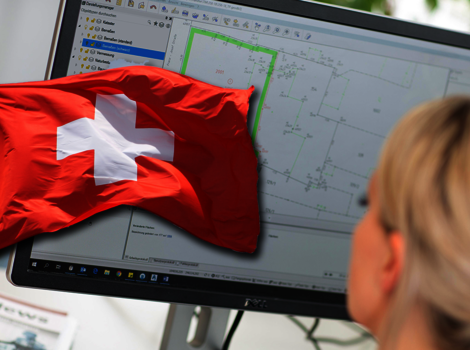 Eine rote Schweizer Flagge mit weißem Kreuz liegt vor einem Computerbildschirm. Auf dem Monitor sind technische Zeichnungen bzw. Diagramme sichtbar. Eine Person mit hellen Haaren ist von hinten zu sehen und blickt auf den Bildschirm.