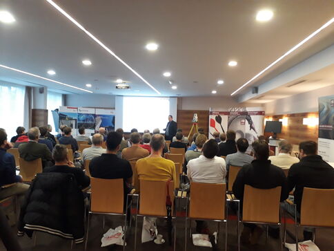 Vortrag in einem Konferenzsaal: Ein Redner steht am Pult vor einer großen Projektion, während etwa zwei Dutzend Zuhörer in Reihen sitzen. Banner und Infotafeln stehen am Rand.