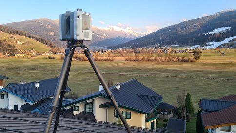Ein Vermessungsgerät auf einem dreibeinigen Stativ steht über einem Dorf mit Häusern, Feldern und Bergen im Hintergrund bei sonnigem Himmel.