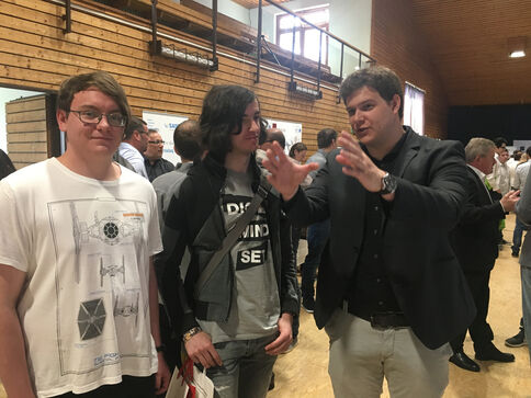 Drei Männer stehen in einem hellen Veranstaltungsraum mit Holzwänden; der Mann rechts gestikuliert erklärend, die beiden anderen hören aufmerksam zu, während im Hintergrund weitere Besucher sprechen.