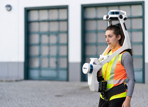 Eine Frau in leuchtender Sicherheitsweste steht draußen vor einem Gebäude und trägt ein großes, weißes Robotik-Exoskelett, das Schultern und Arme stützt; ein Kopfgerät ist mit dem Anzug verbunden.