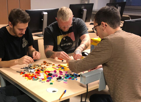 Drei Männer sitzen an einem Tisch und sortieren bunte Kunststoffbausteine, um gemeinsam ein Mosaik zu legen. Auf dem Tisch liegen viele farbige Teile, im Hintergrund sind Computerbildschirme sichtbar.