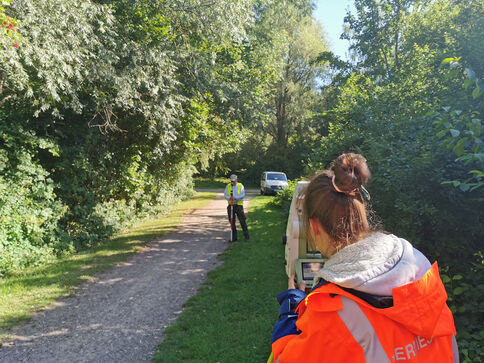 Auf einem von Bäumen gesäumten Kiesweg steht eine Person in einer Warnweste in der Mitte des Weges; eine weitere Person in einer orangefarbenen Jacke fotografiert sie. Im Hintergrund steht ein weißes Auto am Weg.