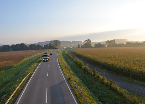 Zweispurige Landstraße durch eine weite Felderlandschaft. Ein Auto fährt von der Kamera weg. Links und rechts Felder mit Hecken und Bäumen, am Horizont sanfte Hügel unter hellem Himmel.