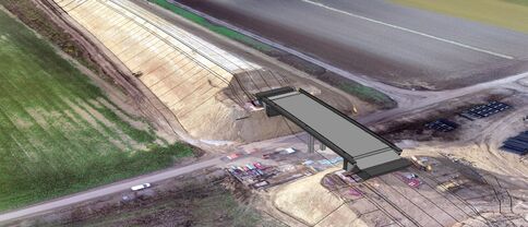 Auf einer Baustelle wird eine lange, dunkle Fußgängerbrücke über Gleise und eine Straße gelegt. Die Brücke liegt schräg über Erdarbeiten, umgeben von Baumaschinen und Bauzubehör.