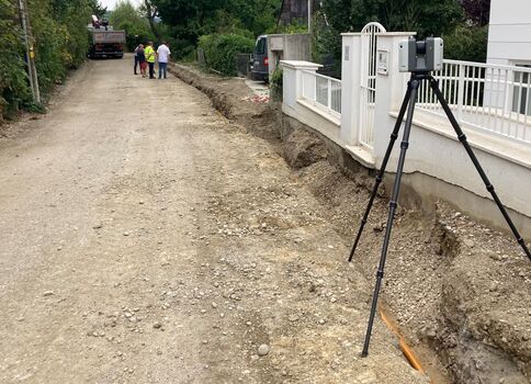 Eine unbefestigte, kiesige Straßenbaustelle. Rechts verläuft ein Graben neben einem weißen Zaun. Ein Stativ mit Vermessungsgerät steht am Rand. Im Hintergrund gehen zwei Personen, und ein Fahrzeug steht weiter. Die Szene zeigt Bau- bzw. Vermessungsarbeiten an der Straße.
