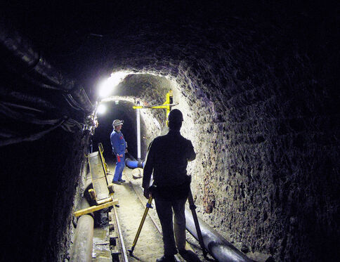 Zwei Bauarbeiter arbeiten in einem dunklen Tunnel; einer steht im Vordergrund mit Helm, der andere weiter hinten neben Geländer und Rohren.