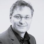 Porträt eines lächelnden Mannes mit Brille und Anzug, der freundlich in die Kamera blickt, vor hellem Hintergrund.