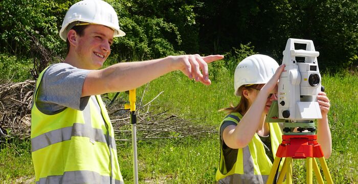Zwei Bauarbeiter tragen Helme und Warnwesten im Freien. Einer zeigt mit dem Arm, der andere bedient eine Vermessungsstation auf einem Stativ; sie führen eine Vermessung im Grünen durch.