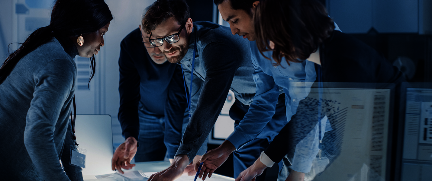 Vier Personen versammeln sich um einen Tisch, schauen auf Unterlagen und arbeiten konzentriert zusammen. Es wirkt wie eine Team-Besprechung in einem Büro.