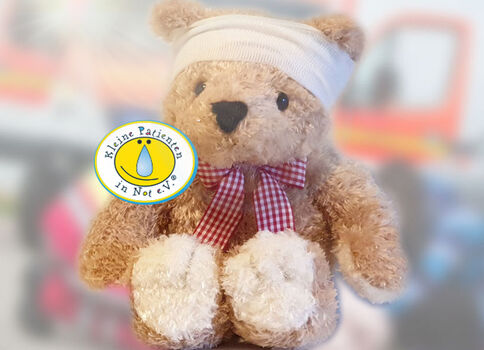 Ein plüschiger Teddybär mit weißem Kopfverband und rotem kariertem Band um den Hals sitzt frontal; auf der Brust trägt er ein rundes Logo der Organisation "Kleine Patienten in Not e.V.".