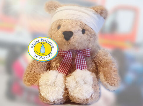 Ein plüschiger Teddybär mit weißem Kopfverband und rotem kariertem Band um den Hals sitzt frontal; auf der Brust trägt er ein rundes Logo der Organisation "Kleine Patienten in Not e.V.".