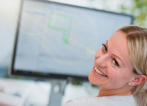 Eine lächelnde Frau sitzt am Arbeitsplatz; im Hintergrund sieht man einen Computerbildschirm mit einer Grafik.