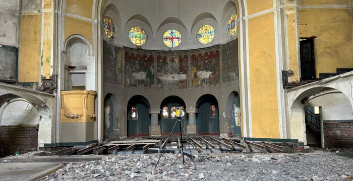 Innenraum einer zerstörten Kirche: trümmerbedeckter Boden, bröckelnde Stufen, bunte Bleiglasfenster und Fresken im Chor, gelbe Wände mit Bögen. Vorne ein Stativ mit Messgerät; der Raum wirkt leer und ruinös.