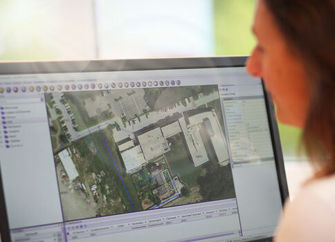 Eine Person betrachtet auf einem Monitor eine Kartenansicht: eine Satellitenaufnahme eines Gebäudekomplexes mit umliegenden Straßen in einer Karten- oder Planungssoftware.