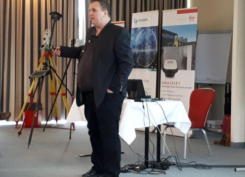 Ein Mann in dunklem Anzug hält einen Vortrag in einem Konferenzraum. Im Hintergrund stehen Banner; vorne rechts ein Tisch mit Laptop und Kabeln, und vor dem Mann steht ein Stativ mit Vermessungsgerät.