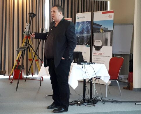 Ein Mann in dunklem Anzug hält einen Vortrag in einem Konferenzraum. Im Hintergrund stehen Banner; vorne rechts ein Tisch mit Laptop und Kabeln, und vor dem Mann steht ein Stativ mit Vermessungsgerät.