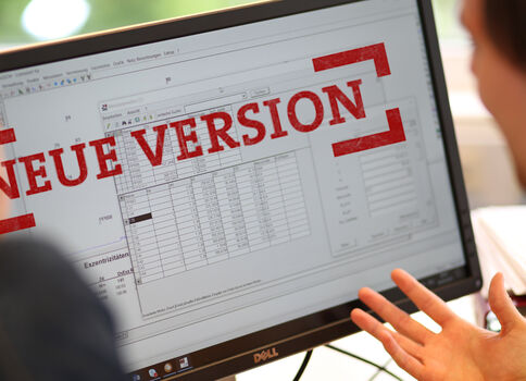 Zwei Personen schauen auf einen Computerbildschirm, der eine Tabellen- bzw. Spreadsheet-Ansicht zeigt. Ein roter Stempel mit der Aufschrift "NEUE VERSION" liegt darüber und weist auf eine neue Softwareversion hin.