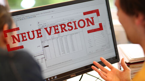 Zwei Personen schauen auf einen Computerbildschirm, der eine Tabellen- bzw. Spreadsheet-Ansicht zeigt. Ein roter Stempel mit der Aufschrift "NEUE VERSION" liegt darüber und weist auf eine neue Softwareversion hin.