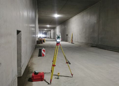 Vue intérieure d’un tunnel en construction. Devant, un niveau laser monté sur un trépied jaune et une mallette rouge. Plus loin, panneaux et cônes de sécurité, éclairage au plafond, murs et sol en béton, et une petite machine de chantier au loin.