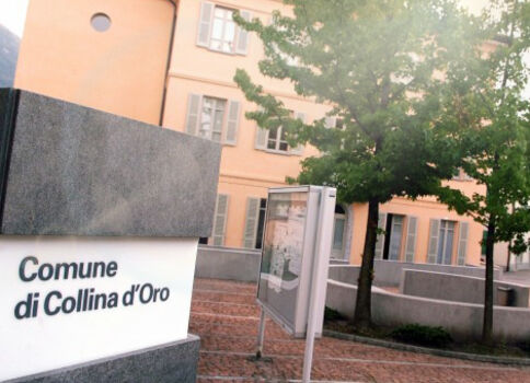 Schild mit der Aufschrift "Comune di Collina d'Oro" vor einem hellorangefarbenen Gebäudekomplex und Bäumen.