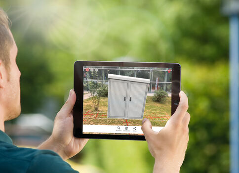 Eine Person hält ein Tablet und fotografiert draußen einen grauen Metall-Schuppen in einem umzäunten Gelände.
