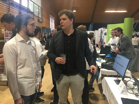 Auf einer Messe sprechen zwei Männer miteinander: Einer in hellem Hemd mit Brille, der andere in dunkler Jacke; der Mann rechts hält ein Getränk. Im Hintergrund sieht man Messestände und weitere Besucher.