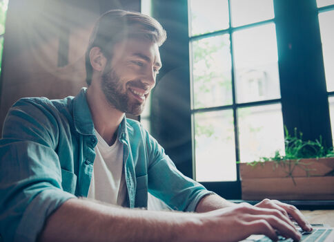 Ein lächelnder Mann mit Bart sitzt an einem Tisch und tippt auf einem Laptop. Er trägt ein blaues Denimhemd über einem weißen T‑Shirt. Helles Tageslicht fällt durch große Fenster; eine Pflanze steht auf der Fensterbank.