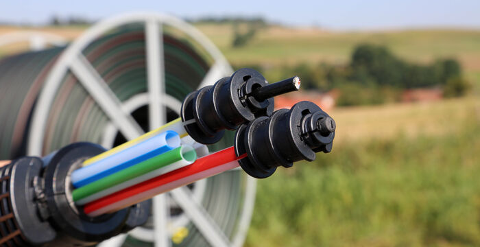 Nahaufnahme eines Bewässerungssystems: Mehrere bunte Schläuche (rot, blau, grün, gelb) schließen sich an schwarze Verbindungsstücke an; dahinter eine Schlauchtrommel und eine offene Felderlandschaft.