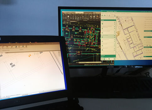 Zwei Computerbildschirme zeigen Baupläne und CAD-Zeichnungen: ein Laptop mit einem Architekturplan und ein größerer Monitor mit einer komplexen CAD-Datei.