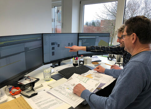Zwei Männer arbeiten an einem Büro-Arbeitsplatz mit drei Monitoren, die Bau- oder Designpläne zeigen. Einer zeigt mit dem Finger auf den Bildschirm, der andere beobachtet. Der Tisch ist voll mit Unterlagen, Skizzen, Notizblöcken und Bürobedarf.