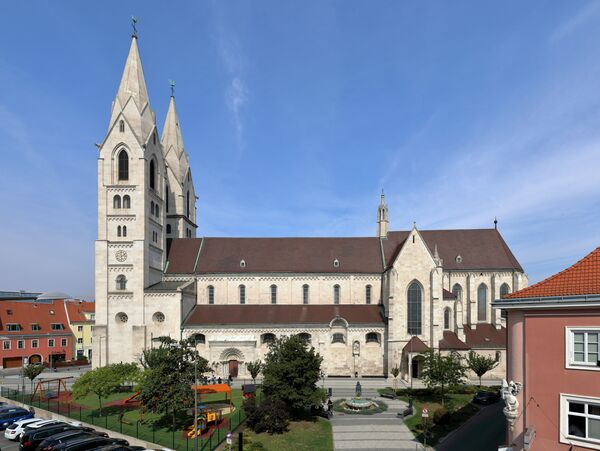 Eine imposante gotische Kirche mit zwei hohen Türmen aus hellem Stein dominiert das Stadtbild; neben ihr erstrecken sich Rotdächer, eine gepflegte Grünanlage mit Wegen und Bäumen sowie einige angrenzende Gebäude.