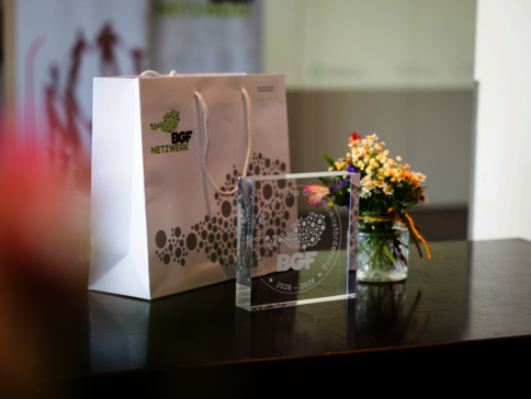 Auf einem dunklen Tisch stehen eine weiße Geschenktüte mit dem BGF Netzwerk-Logo, ein transparentes Acryl-Preisschild mit dem BGF-Logo und Jahreszahlen, sowie ein kleiner Blumenschmuck in einer Glasvase; der Hintergrund ist unscharf.