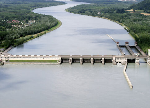 Luftaufnahme eines Flussdamms mit mehreren Toren, der Wasser hinter einer grünen Uferlandschaft staut. Rechts eine Wehranlage, links Gebäude am Flussufer, dahinter Wälder und Felder.