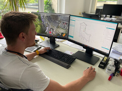 Ein Mann sitzt an einem Schreibtisch mit zwei Monitoren: Der linke Bildschirm zeigt Außenüberwachungsvideos, der rechte Bildschirm eine technische Zeichnung oder Architekturansicht. Er tippt mit der rechten Hand und bedient die Maus, in einem hellen Büro mit Pflanzen.