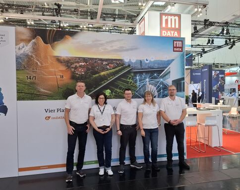 Fünf Personen in weißen Shirts stehen vor dem Messestand der rm DATA. Eine große Panelwand zeigt eine Landschaft mit Feldern und Straßen, überlagert von digitalen Netzstrukturen; im Hintergrund weitere Messestände.
