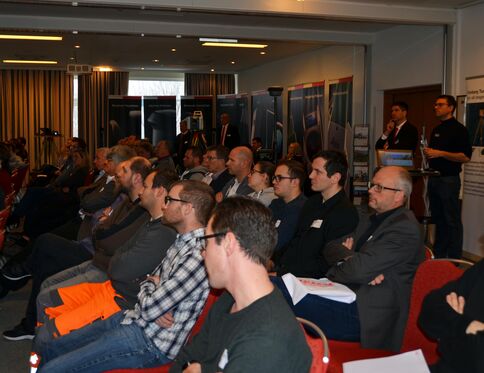 Eine Konferenzsituation: Ein Publikum aus Fachleuten sitzt aufmerksam in einem Konferenzsaal; im Hintergrund stehen Bannerwände und eine Moderationsstation.