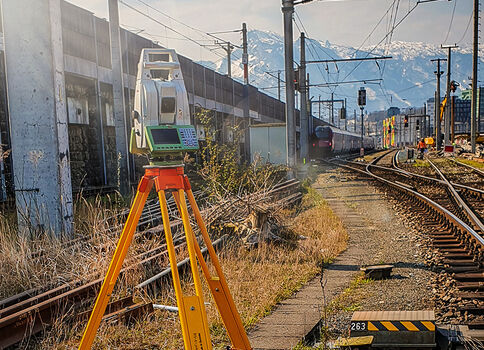 Vermessungsausrüstung: Ein Totalstationsgerät auf einem orangefarbenen Stativ steht neben einer Bahnstrecke. Im Hintergrund erkennt man Züge, Gleise und Überkopfleitungen sowie Berge. Die Aufnahme hebt Messarbeiten im Bahnbereich hervor.