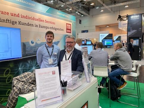 Zwei Männer stehen hinter einem Messestand auf einer Fachmesse. Auf dem Tisch liegen Flyer, Stifte und Visitenkarten; Bildschirme zeigen Diagramme. Im Hintergrund sind weitere Stände und Besucher zu sehen.