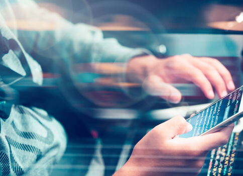 Nahaufnahme: Eine Person hält ein Smartphone, während im Hintergrund eine weitere Person an einer Tastatur arbeitet; beide nutzen digitale Geräte.