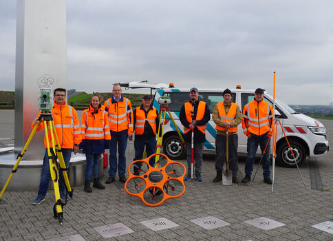 Eine Gruppe Vermessungstechniker in leuchtend orange Warnwesten steht nebeneinander auf einer gepflasterten Fläche; im Hintergrund ein weißer Dienstwagen. Vor ihnen liegen Vermessungsgeräte, darunter Stative und ein orangefarbenes Rad mit Speichen, sowie Markierungen auf dem Boden.