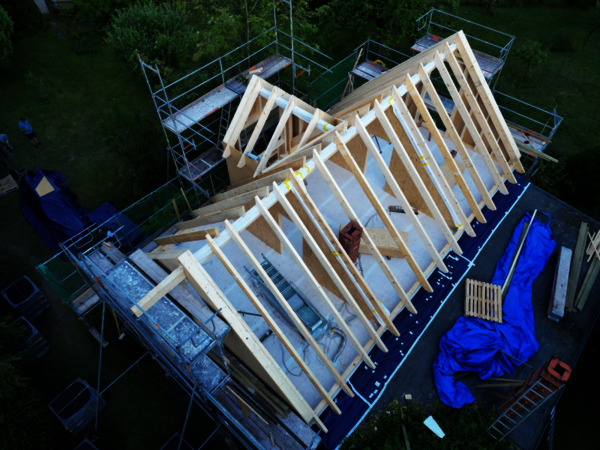 Aerial view of a building under construction with a wooden roof frame formed by triangular trusses, surrounded by scaffolding and blue tarps.