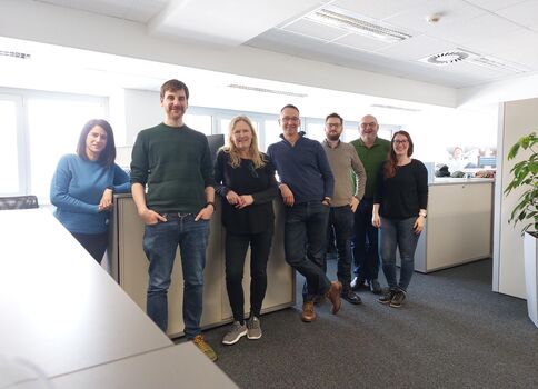 Sieben Kollegen stehen in einem offenen Büro, lächeln zur Kamera, Schulter an Schulter, leger gekleidet; hinter ihnen Schreibtische, Trennwände und große Fenster.