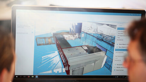 Ein Monitor zeigt ein detailliertes 3D-Modell eines Industrie-Dachkomplexes mit blauen Flächen und Metallstrukturen; im Vordergrund beobachten zwei Personen den Bildschirm.