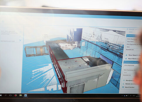 Ein Monitor zeigt ein detailliertes 3D-Modell eines Industrie-Dachkomplexes mit blauen Flächen und Metallstrukturen; im Vordergrund beobachten zwei Personen den Bildschirm.