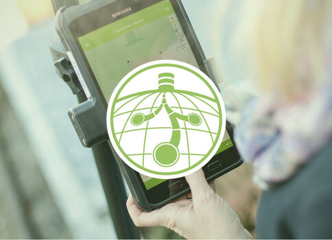 Eine Person hält ein Tablet in einer Halterung; der Bildschirm leuchtet grün, vermutlich mobiles Bezahlen oder Ticketkontrolle. Ein runder, grün-weißer Globus-Logo liegt mittig über dem Gerät.