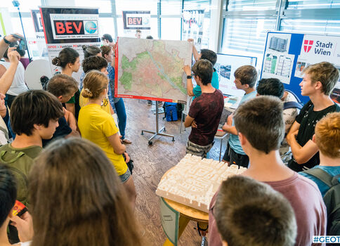 Eine Gruppe junger Menschen steht in einem Ausstellungsraum um eine große Karte, die auf einem mobilen Stativ präsentiert wird; alle schauen aufmerksam darauf und diskutieren offenbar über das gezeigte Thema.