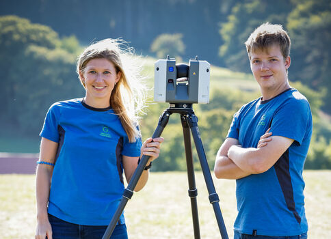Zwei Jugendliche in blauen T-Shirts stehen draußen neben einem Vermessungsgerät auf einem Stativ und vermessen das Gelände.