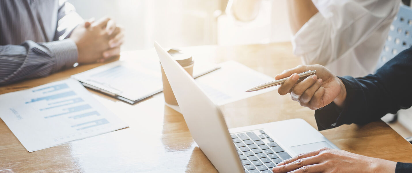 Zwei Personen arbeiten in einer Besprechung an einem Laptop auf einem Tisch. Rechts hält eine Person mit Maske einen Stift und blickt auf den Bildschirm; auf dem Tisch liegen Unterlagen und Diagramme.