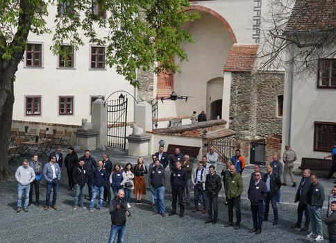 Eine Gruppe von Menschen steht in einem historischen Innenhof vor einer Burg- oder Schlossanlage. Über ihnen fliegt eine Drohne; im Hintergrund sind Torbogen, Steinmauern und Bäume sichtbar.